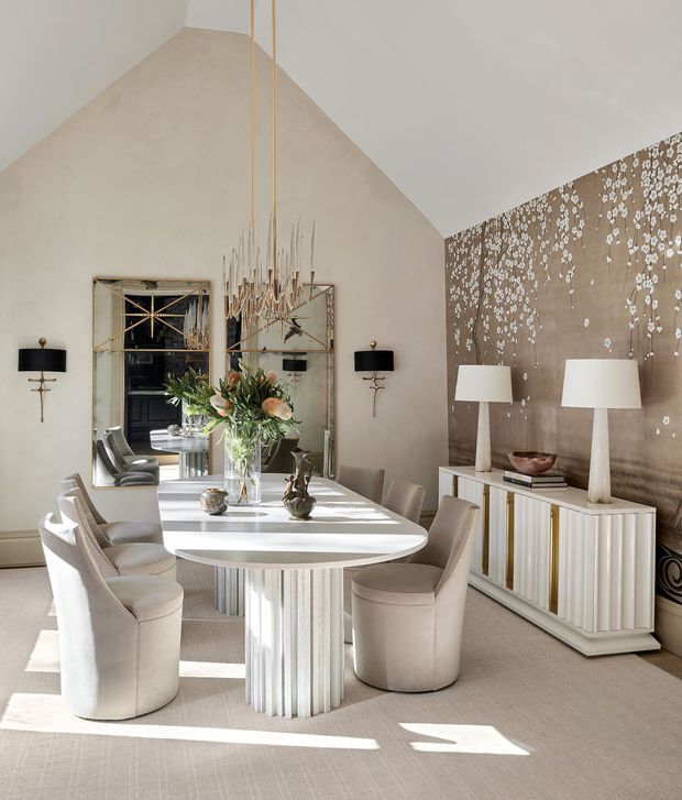 Elegant contemporary dining room with a tall vaulted ceiling, a sculptural oval dining table surrounded by upholstered dining chairs, and a statement chandelier suspended above. The room features warm neutral tones, a textured taupe feature wall with a delicate white botanical pattern, large mirrored panels, a fluted white sideboard, matching table lamps, and abundant natural light.