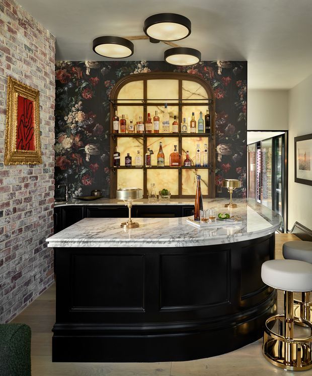 A sophisticated home bar with a curved black paneled island topped in veined marble. Behind it, an arched, backlit display cabinet showcases liquor bottles against dark floral wallpaper. The space features exposed brickwork, framed artwork, brass bar stools and fixtures, and sculptural circular ceiling lights.