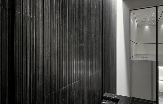 A dramatic monochromatic interior featuring a full-height dark vertically ribbed feature wall beside a glazed opening into a bright minimalist room. The space has sharp geometric lines, recessed ceiling spotlights, dark stone-like flooring, and a restrained high-contrast palette.