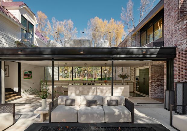A contemporary multi-level courtyard-like living space viewed from outdoors, with a large upholstered sectional sofa in the foreground, an open-plan kitchen and dining area behind it, extensive black-framed glazing, exposed brick walls, and a flat-roofed connection between two building volumes. Mature trees are visible beyond the courtyard, and the space is brightly lit with a warm, refined indoor-outdoor atmosphere.