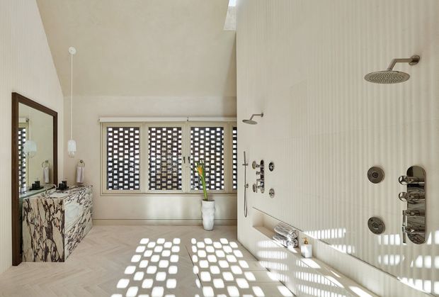 Bright, refined bathroom with a walk-in multi-shower installation, wall-mounted controls, and a freestanding or integrated shower area. The room features pale vertically textured wall surfaces, light herringbone flooring, a marble-patterned floating vanity with dark timber edging, a large framed mirror, and patterned screening over the windows that casts geometric sunlight across the floor and walls.