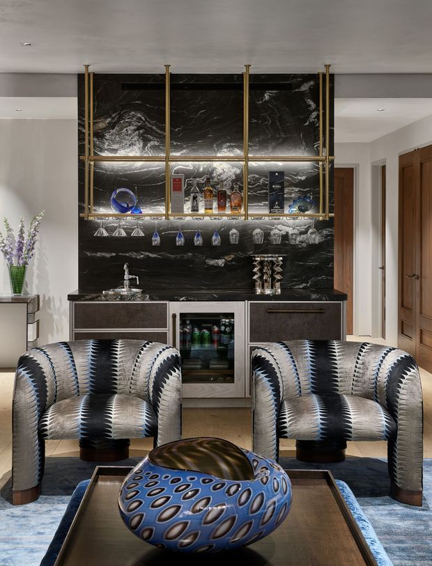 A sophisticated home bar or lounge area centered on a dramatic black marble feature wall and illuminated brass-framed glass shelving. The shelving displays bottles and glassware above a compact bar counter with sink, cabinetry, and under-counter beverage refrigerator. Two patterned armchairs sit in the foreground around a dark coffee table with a sculptural blue glass vessel. The room combines glossy dark stone, warm timber doors, brass detailing, and bold geometric upholstery in a luxurious, dramatic interior.