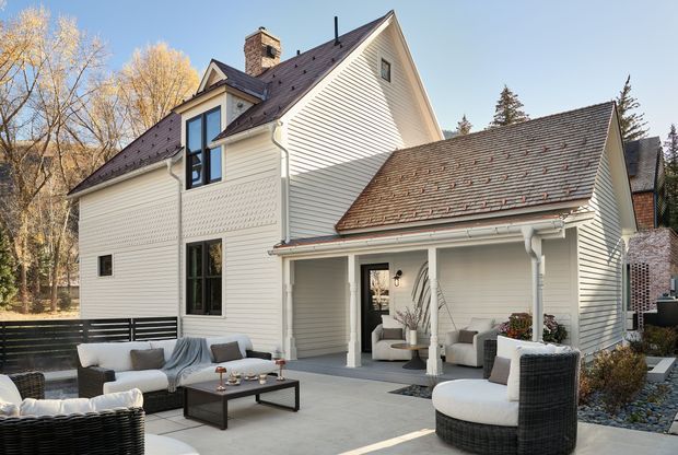 Exterior view of a substantial multi-gabled house with pale horizontal weatherboard cladding, dark pitched roofs, a brick chimney, and a covered outdoor living area. The landscaped terrace includes outdoor seating and a low coffee table, with mature trees surrounding the property.