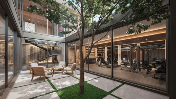 A refined contemporary courtyard interior with a mature tree planted in a square lawn bed, surrounded by large-format pale paving slabs and narrow green joints. Floor-to-ceiling glazing connects the courtyard to a luxurious home gym with exercise equipment, timber flooring, warm recessed and linear lighting, and built-in display or storage joinery. A lounge area with timber-framed armchairs and a low table is visible on the left, while a glazed upper level and timber-lined soffits create a layered indoor-outdoor composition.