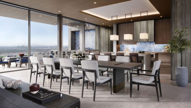 A spacious, refined open-plan dining and kitchen interior with floor-to-ceiling glazing overlooking a distant cityscape. A large dark dining table is surrounded by upholstered chairs, while the rear kitchen features dark timber cabinetry, a pale stone benchtop, and a blue-toned patterned splashback. Vertical timber-textured wall panels, a slatted timber ceiling, pendant lights, and a substantial structural column create a warm contemporary setting.