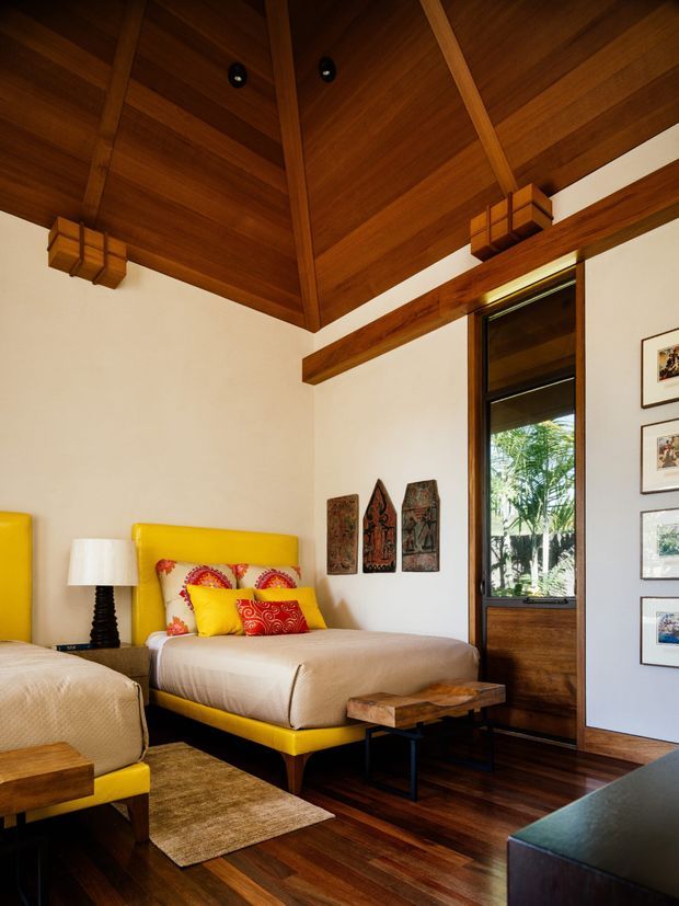 Warm, richly timber-lined bedroom with a dramatic exposed pitched ceiling and structural beams. Two yellow upholstered beds with colourful patterned cushions sit on dark timber flooring, accompanied by a bedside lamp, woven rug, and rustic timber coffee tables. A tall timber-framed glazed door or window opens to greenery outside, while framed artwork decorates the walls.
