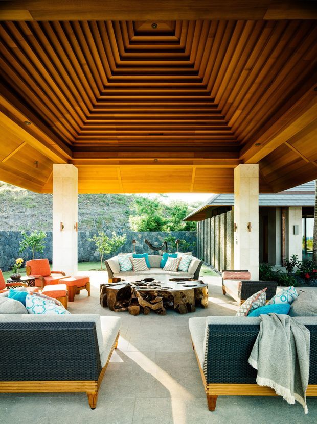 A spacious covered outdoor lounge with a dramatic recessed, tiered timber ceiling and exposed wood beams. The seating area includes woven lounge chairs and sofas with turquoise and coral cushions, arranged around a substantial sculptural natural-wood coffee table. Pale stone or concrete flooring, square stone-clad columns, and an open view toward a landscaped garden and adjacent building create a warm, resort-like indoor-outdoor setting.
