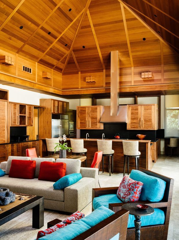 A spacious open-plan living, dining, and kitchen area beneath a dramatic timber-lined vaulted ceiling. The room features extensive warm wood cabinetry and wall panelling, a large kitchen island with upholstered bar stools, black splashback and rangehood, and a seating area with a neutral sectional sofa and vivid turquoise, red, and patterned cushions. Exposed timber framing and integrated ceiling spotlights create a strong lodge-like architectural character.