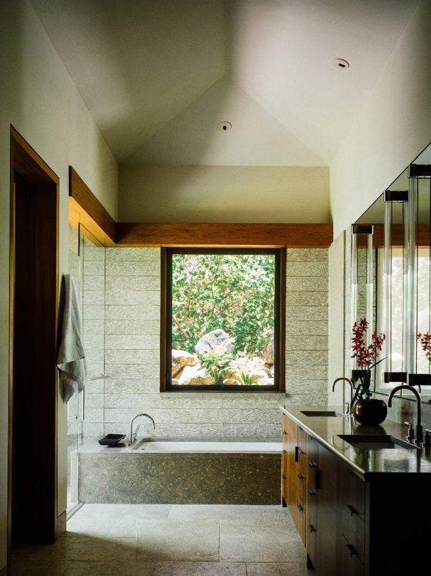 A refined bathroom viewed from the entry, featuring a freestanding stone bath beneath a large garden-facing window, pale textured stone wall tiles, large stone floor tiles, dark timber framing and cabinetry, and a long stone vanity with twin black taps and mirrors. A glass shower enclosure is visible on the left, while the ceiling has angled geometric planes and recessed lighting.