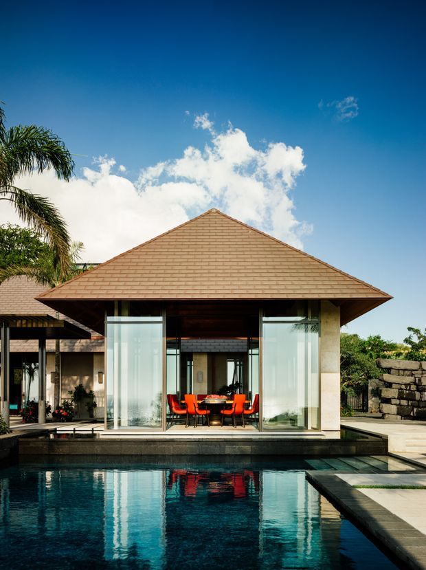 A tropical, resort-like house viewed across a rectangular swimming pool. The main pavilion has a broad pyramidal terracotta-tiled roof, slender stone-clad supports, and extensive floor-to-ceiling glazing. Inside, a dining table with vivid orange-red chairs is visible, while additional tiled-roof structures, tropical planting, and a dark stone boundary wall frame the scene.