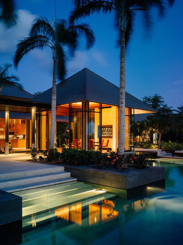 Twilight view of a luxurious tropical modern residence opening onto a landscaped swimming pool. The pavilion-like building has broad low-pitched hipped roofs with dark horizontal cladding, extensive floor-to-ceiling glazing, warm illuminated interiors, timber-lined ceilings, and generous stone steps leading into the pool. Tall palm trees and planted garden beds frame the architecture.