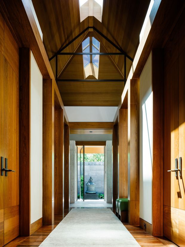 A dramatic residential hallway lined with rich timber wall panelling and doors, featuring a long light-coloured runner, strong axial views, and a vaulted timber ceiling. Black steel trusses frame a central skylight, while clerestory-like glazing and tall side windows bring in bright natural light. The corridor terminates at a glazed opening toward a small landscaped courtyard with a water feature.