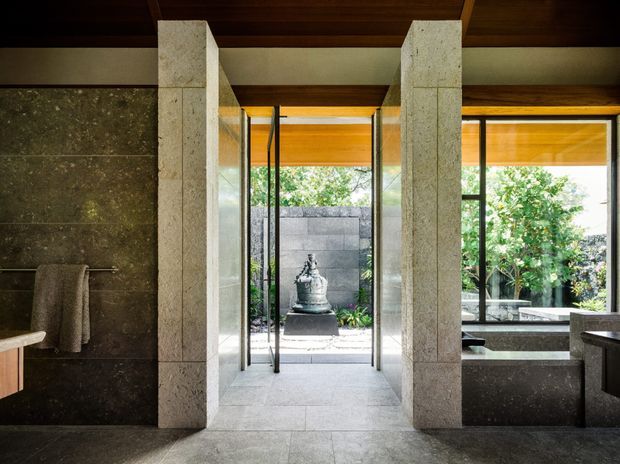 A refined, dark-toned bathroom interior with large-format stone wall and floor finishes, a freestanding or partially visible bath area to the right, and a timber ceiling with exposed beams. Two substantial pale stone piers frame full-height glazing and a central opening toward a landscaped courtyard with a sculptural water feature. The room combines strong indoor–outdoor connection, natural materials, and high-contrast architectural detailing.
