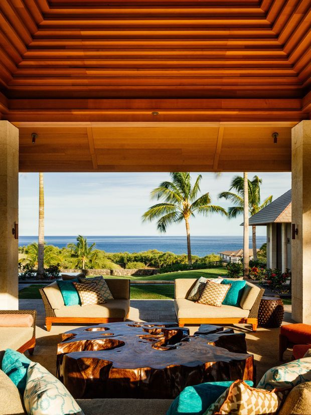 A luxurious open-sided living room or outdoor lounge overlooking a tropical ocean coastline. The space features a richly detailed dark timber coffered ceiling, stone columns, large upholstered sofas with turquoise and patterned cushions, and a substantial organic live-edge timber coffee table. Palm trees, landscaped grounds, and the ocean are visible beyond the covered terrace.