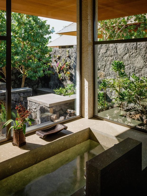 A tranquil indoor-outdoor bathing space with a sunken rectangular water basin or bath beside floor-to-ceiling corner glazing. Warm timber ceiling, dark metal window frames, textured stone walls, and a landscaped courtyard with rough stone seating and tropical planting create a natural, resort-like atmosphere.