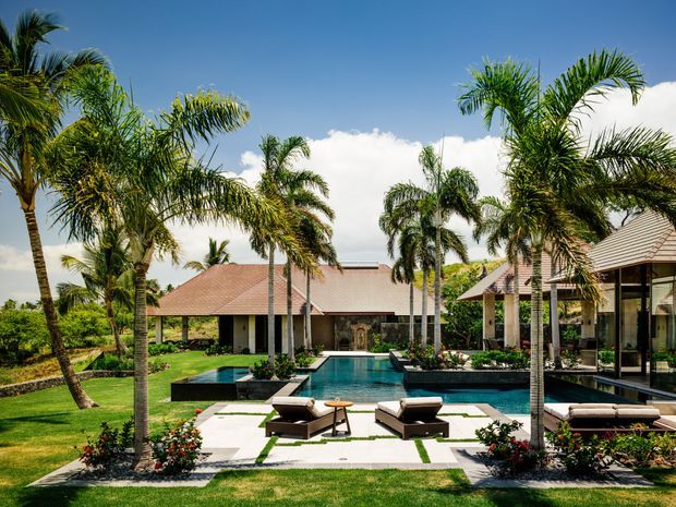 A tropical resort-style residential property viewed from a landscaped garden, with a rectangular swimming pool, paved sun deck, loungers, palm trees, planted garden beds, and low-rise buildings with terracotta tiled roofs and extensive glazing.