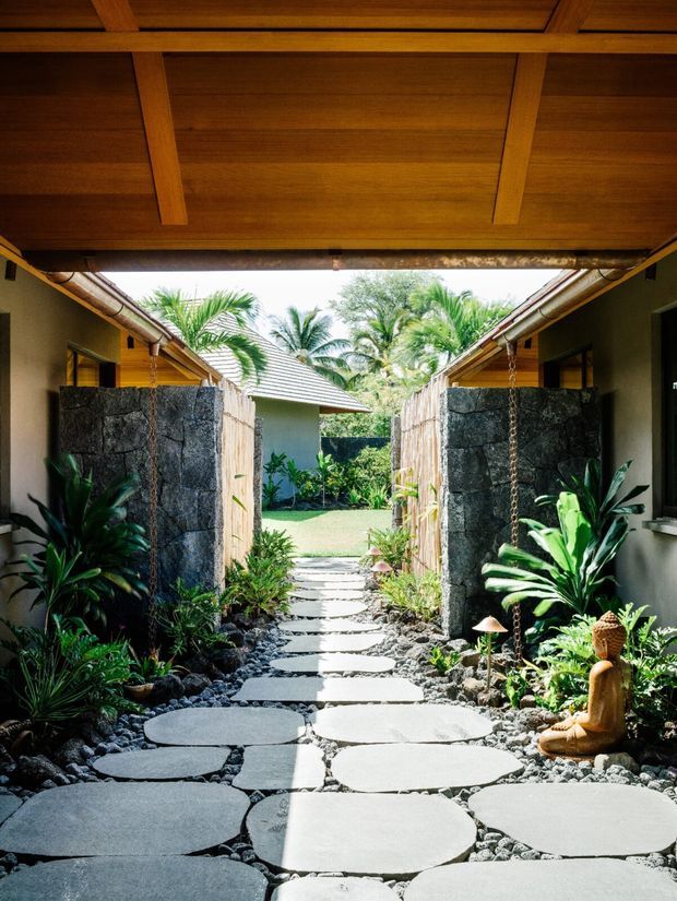 A tropical landscaped courtyard walkway viewed beneath a timber-clad overhead canopy. Large irregular pale stone stepping slabs run through dark pebbles and lush planting, with textured dark stone walls, rain chains, timber fencing, and low pavilion-like buildings on either side. The path opens toward a sunny garden beyond, creating a sheltered, resort-like outdoor passage.