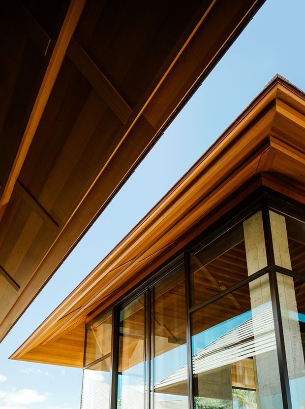 Upward architectural detail of a contemporary building with extensive warm timber soffits and deep roof overhangs, dark-framed floor-to-ceiling glazing, and exposed concrete structural columns. The composition emphasizes layered rooflines, timber construction, and crisp geometric junctions against a clear blue sky.