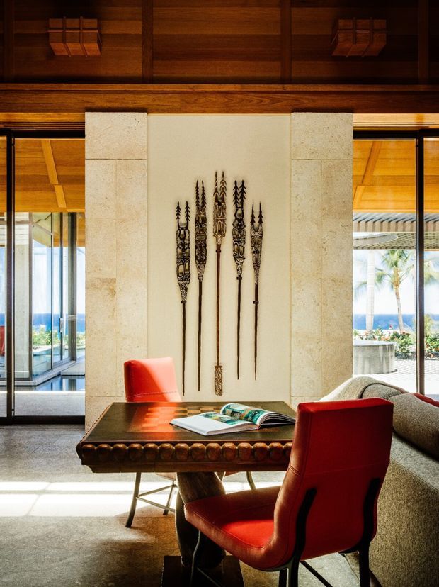 A richly detailed interior lounge or study area with a dark timber ceiling and beams, warm wood wall panels, pale stone wall cladding, and large glazed openings overlooking a bright coastal setting. A substantial carved dark-wood table with books is paired with vivid red leather chairs and a curved upholstered sofa. Decorative carved spears are mounted on the central wall, adding an eclectic artisanal character.