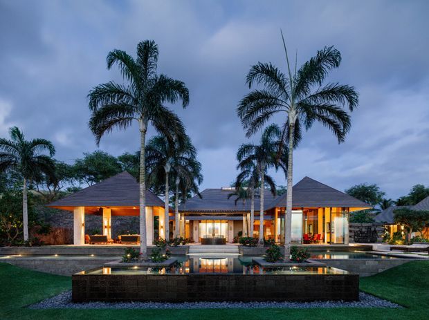 A symmetrical luxury resort-style residence viewed from the landscaped front garden at dusk. The low, pavilion-like building has multiple dark hipped roof forms, broad open glazing, warm illuminated interiors, pale structural columns, and a long reflecting pool with raised stone edging in the foreground. Tall palm trees and manicured lawns frame the composition.