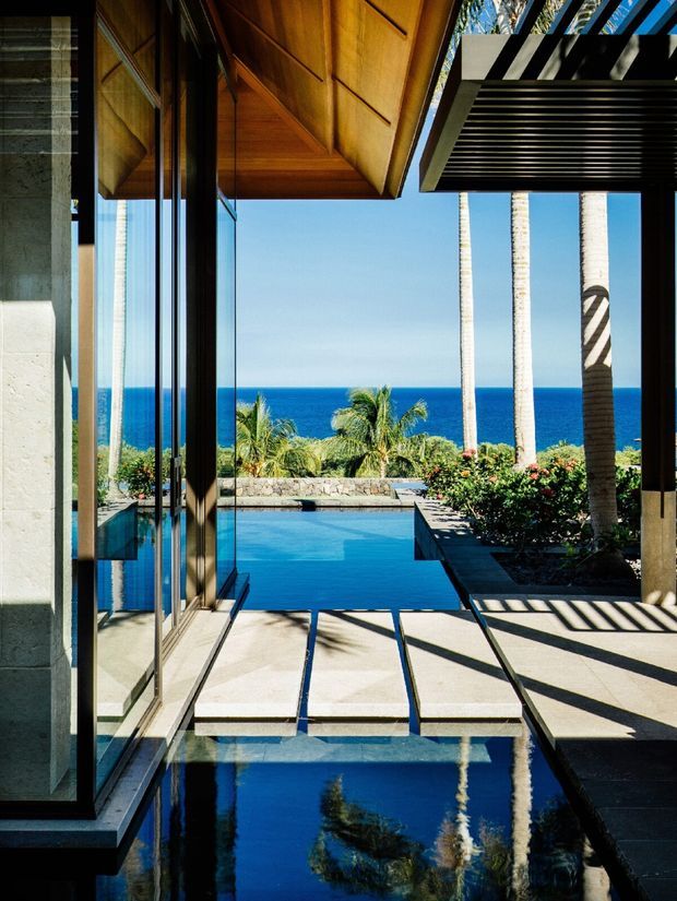A dramatic architectural view from a glazed indoor space toward a swimming pool and ocean beyond. Full-height glass doors frame a covered terrace with a timber-lined projecting roof and bold slatted overhead structure. Pale stone paving, dark structural elements, tropical palms, and strong reflections in the water create a luxurious coastal indoor-outdoor connection.