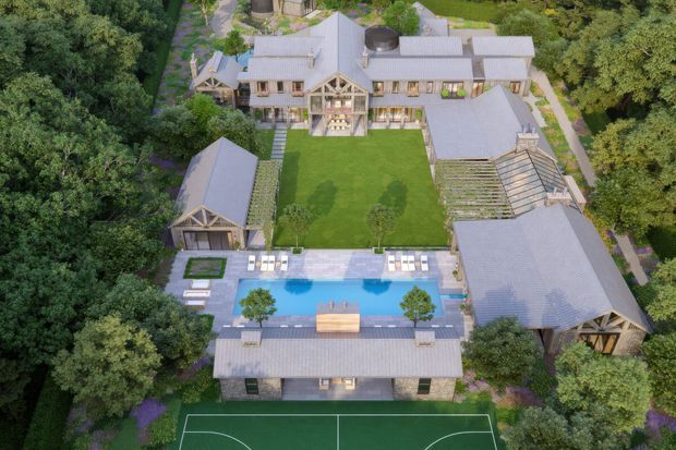Aerial view of a large landscaped residential estate surrounded by mature trees. The property comprises several pitched-roof buildings arranged around a central lawn, with a swimming pool and paved terrace, a sports court, and extensive garden paths and planting. The architecture has a refined traditional country-estate character with light-coloured rendered walls, timber detailing, and pale metal roofing.