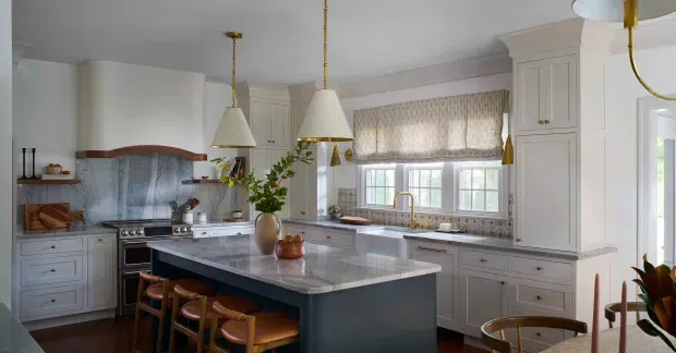 A refined traditional kitchen with pale built-in cabinetry, a large central island, stainless steel range, patterned blue-grey backsplash, marble-look benchtops, pendant lighting, and warm timber stools. A bay-style window with a fabric Roman blind provides natural light.