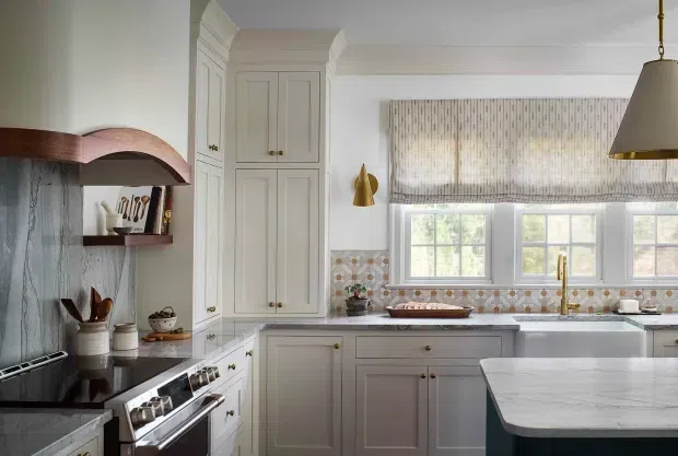 A refined traditional kitchen with white painted cabinetry, a dark range and benchtop, patterned tile splashback, marble-look island, farmhouse sink, brass fixtures, and a window lined with patterned roman blinds. Decorative arched timber shelving and detailed cabinetry add character.