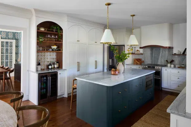 A refined traditional kitchen with a large deep blue-green island, pale stone benchtops, white cabinetry with arched detailing, brass hardware, timber flooring, and matching pendant lights. The space includes a built-in drinks station with open timber shelving, a patterned tiled splashback, rangehood, and a small dining area by the glazed door.
