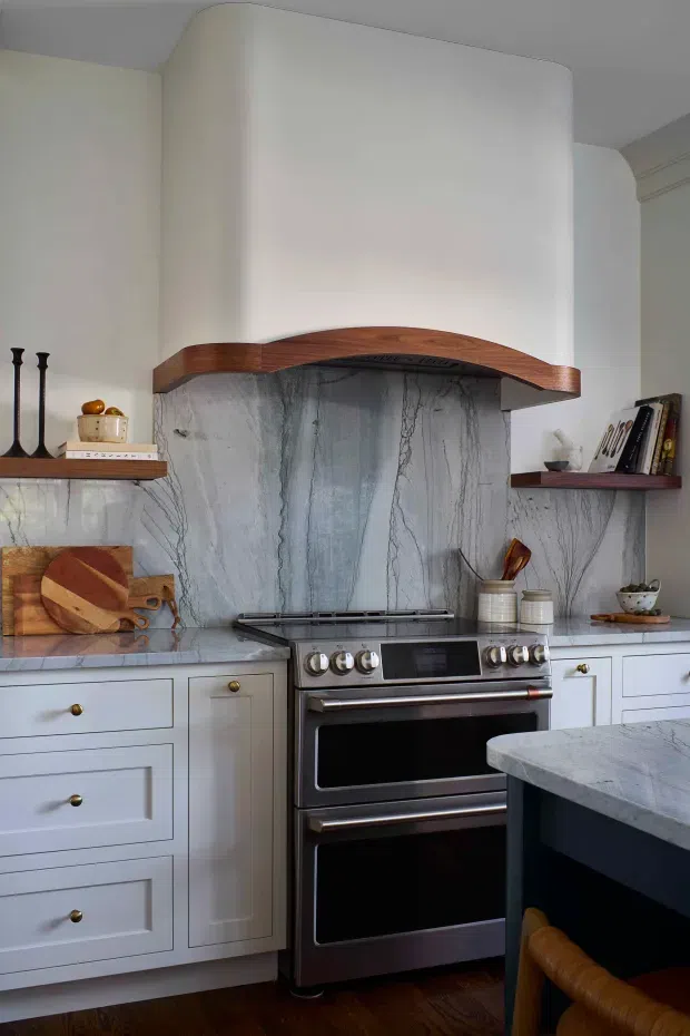 A refined kitchen centered on a large stainless-steel range beneath a sculptural curved plaster range hood. Veined grey marble forms the backsplash and benchtops, contrasting with pale painted cabinetry, brass knobs, dark timber open shelving, and a dark island. The room has a warm, classic character with carefully detailed joinery and natural materials.