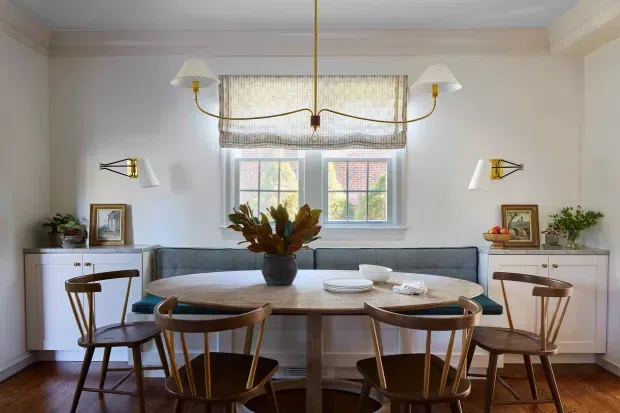 A refined dining room with a built-in upholstered banquette beneath a window, a large round timber dining table, and six curved wooden dining chairs. Symmetrical white built-in cabinetry flanks the banquette, with framed artwork, plants, tableware, and warm brass wall and pendant lighting. The room features pale walls, timber flooring, classic mouldings, and a restrained contemporary-traditional aesthetic.