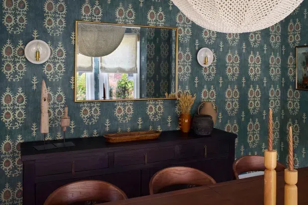 A richly decorated dining area featuring dark patterned botanical wallpaper, a long dark timber sideboard, a large brass-framed mirror, sculptural wall sconces, ceramic vessels, and warm timber dining chairs and candlesticks. The room has an eclectic, moody and refined character with layered vintage-inspired details.