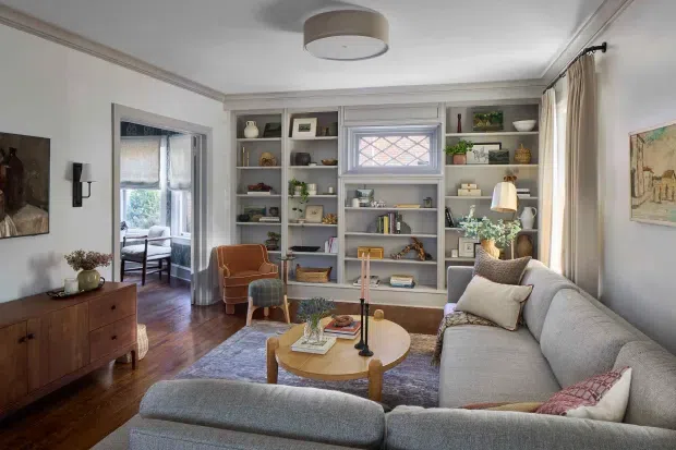 A warmly styled living room with a large light-grey sectional sofa, rounded timber coffee table, patterned area rug, and mid-century-inspired armchair. Built-in grey shelving surrounds a small decorative window and displays books, ceramics, plants, and artwork. The room has white walls and ceiling with traditional cornice detailing, dark timber flooring, cream curtains, a timber sideboard, and layered ambient lighting. An adjoining glazed sitting area is visible through the opening on the left.