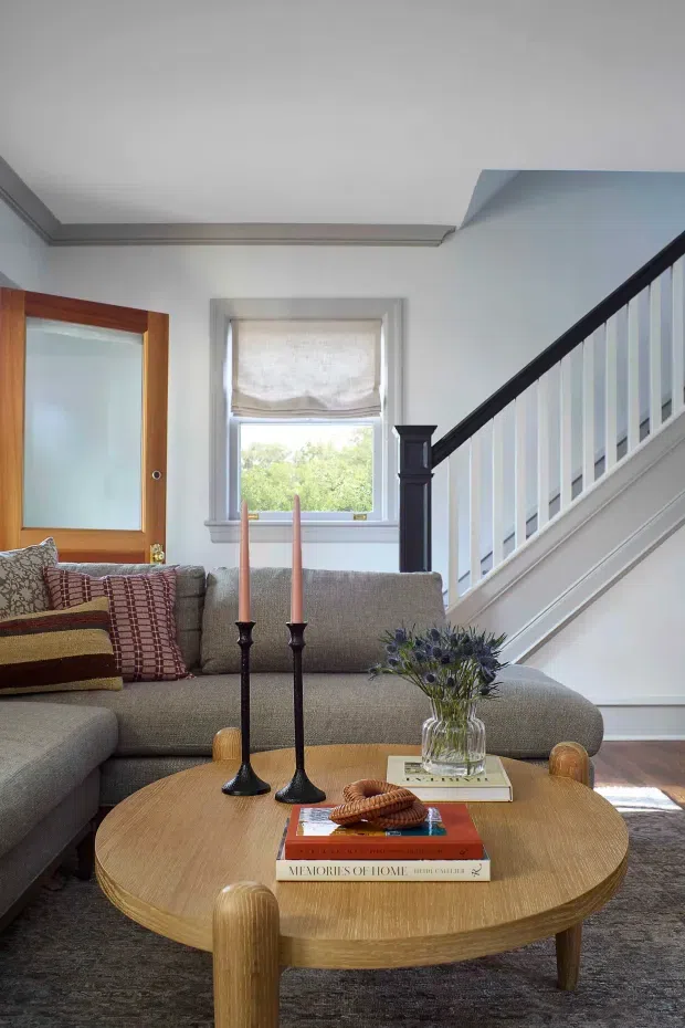 A bright, traditionally detailed living room with a large grey sectional sofa, patterned cushions, and a round light-wood coffee table styled with books, candlesticks, and a vase of flowers. A timber-framed glazed door, upholstered window shade, decorative ceiling trim, and a staircase with dark timber handrail and white balusters are visible.
