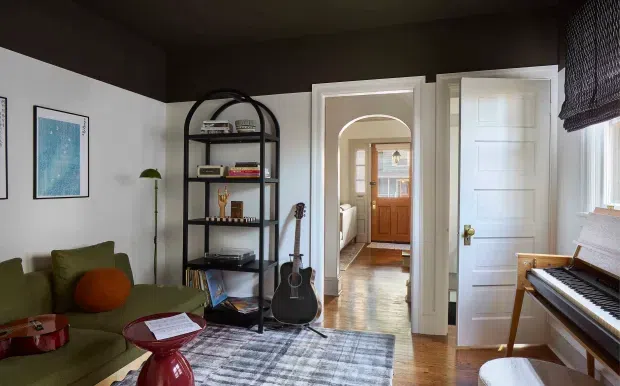 A warm, eclectic living room with a green sofa, arched black metal shelving, guitar, framed artwork, patterned rug, and upright piano. Dark painted walls and ceiling contrast with white trim, while an arched doorway leads through to a timber-floored hall and front door.