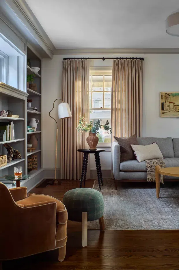 A warm, traditionally detailed living room with built-in open shelving, a large window dressed in full-length beige curtains, a grey sofa, leather armchairs, a patterned ottoman, a small dark wood side table, and a soft area rug. Dark timber flooring, decorative wall and ceiling mouldings, a curved floor lamp, and layered furnishings create a refined, cozy atmosphere.