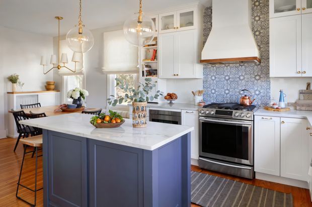 Bright, refined kitchen and dining space with a large navy-blue island topped in white stone, white shaker-style cabinetry, patterned blue-and-white tile backsplash, stainless-steel range, and a sculptural white range hood. Brass pendant lights and a brass dining chandelier add warmth, while timber flooring, a striped runner, floral arrangements, and a compact dining area create a classic yet fresh atmosphere.