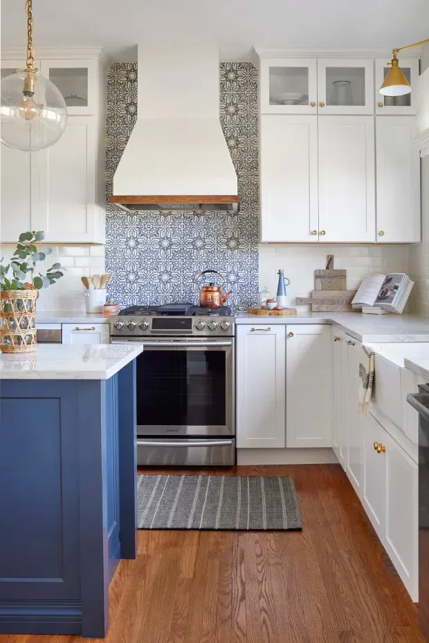 A bright, compact kitchen with white shaker-style cabinetry, a deep blue island, stainless steel range and appliances, patterned blue-and-white tile backsplash, white subway-tile side walls, and warm timber flooring. Brass hardware and pendant lighting add warmth, while open upper cabinet sections display simple glassware.