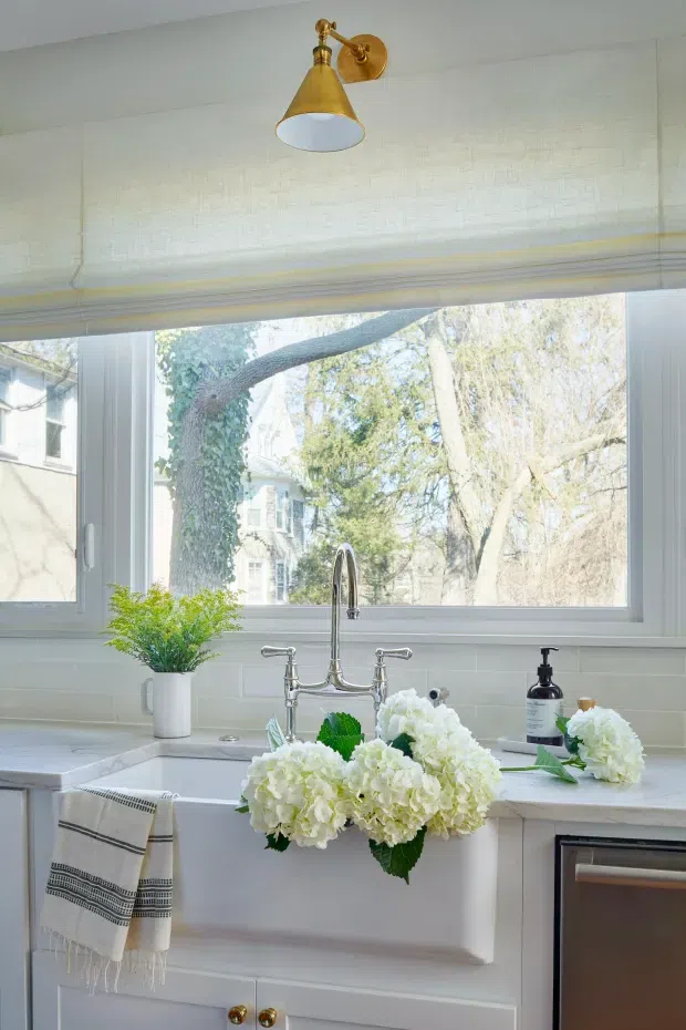 Bright, refined kitchen sink area beneath a large window. The space features white tiled walls, pale cabinetry, a light stone or composite countertop, a deep farmhouse-style sink with a polished metal bridge faucet, and a brass wall sconce. White roller blinds, a striped tea towel, soap dispenser, greenery, and white flower arrangements complete the styled scene. Mature trees and neighbouring architecture are visible through the window.