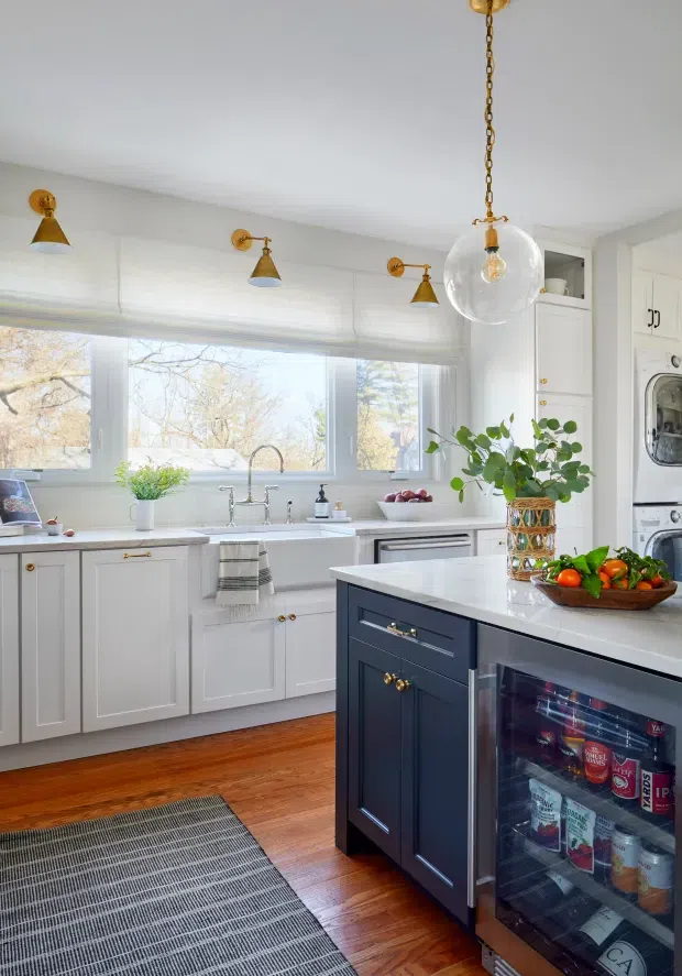 A bright, refined kitchen with white perimeter cabinetry, a dark navy-blue island, white stone benchtops, timber flooring, large windows with roman blinds, brass hardware, and pendant lighting. The island includes a glass-front beverage or wine refrigerator, while fresh greenery and produce add colour.
