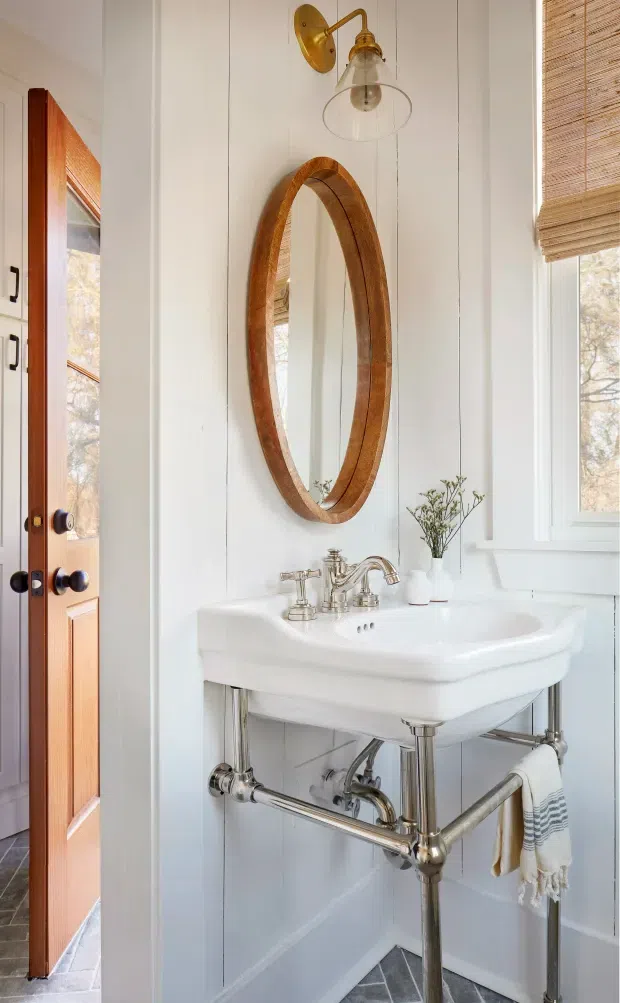 A compact, bright bathroom with a white pedestal-style washbasin and exposed polished chrome plumbing, set against white vertical wall panelling. A tall oval timber-framed mirror hangs above the basin, paired with a brass wall sconce and clear glass shade. A timber door, window with woven bamboo blind, small vase, and folded towels add warm natural accents. Grey geometric floor tiles are visible below.