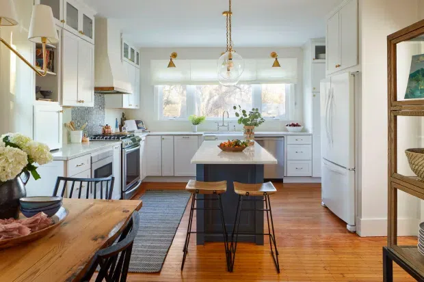 Bright, traditionally detailed white kitchen with a central island, integrated cabinetry, stainless steel range and refrigerator, patterned backsplash, timber flooring, and a connected dining area. Natural light enters through a wide window above the rear benchtop, while brass and glass pendant lighting adds warmth.