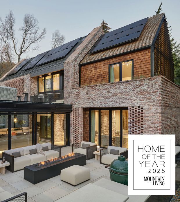 Exterior view of a substantial multi-level home arranged around a paved courtyard patio. The building combines weathered red brick walls, timber shingle-clad gabled roof sections with extensive solar panels, dark-framed glazing, and a contemporary glazed extension. The courtyard contains upholstered outdoor seating, a long linear fire feature, and a green ceramic grill. A visible graphic reads “Home of the Year 2025 Mountain Living.”