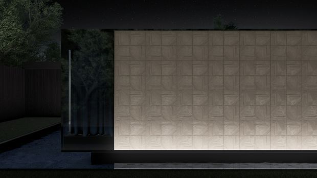 Night-time exterior view of a minimalist, flat-roofed building facade. A large illuminated wall is clad in repeating textured rectangular panels, bordered by dark horizontal bands and dark glazing. Trees, a fence, and a paved approach are visible around the structure.