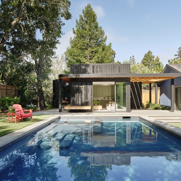 A contemporary backyard pool area with a compact dark-clad pavilion or pool house beside a larger residence. The pavilion has a flat roof, deep timber eaves, extensive glazing, and an open covered connection to the main building. A rectangular swimming pool dominates the foreground, with pale paving, mature trees, timber fencing, and red outdoor chairs contributing to a relaxed landscaped setting.