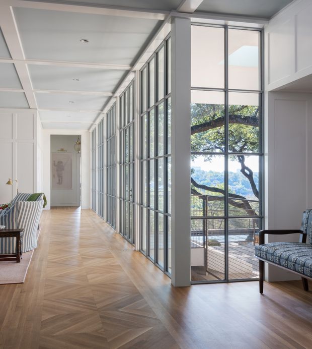 A bright, refined interior hallway or living circulation space with a long wall of black-framed, multi-pane glazed doors and windows opening to an elevated outdoor terrace. The room features pale white walls and ceiling beams, light herringbone timber flooring, upholstered striped seating on the left, and a patterned armchair on the right. The glazing provides strong views of mature trees and a distant landscape, creating an airy connection between interior and exterior.