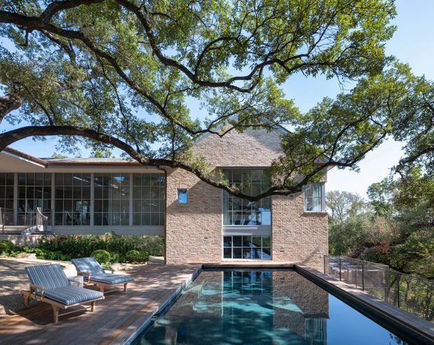 Exterior view of a substantial brick-clad residence opening onto a tiled pool terrace. Large glazed living areas sit beside a taller central brick volume, while mature spreading trees frame the building and pool. Outdoor lounge chairs, pool fencing and landscaped grounds are visible.