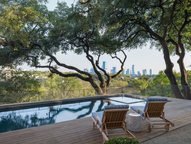 A landscaped outdoor pool terrace surrounded by mature, spreading trees, with a timber deck, glass or wire safety railing, and two wooden reclining lounge chairs beside the pool. A distant modern city skyline is visible beyond the tree canopy.