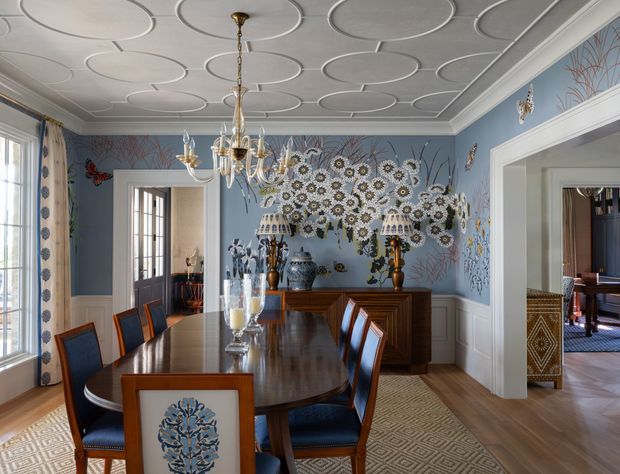 A formal dining room with a large dark timber oval dining table and upholstered blue dining chairs. The room features pale blue walls with white wainscoting and trim, an elaborate floral mural, patterned area rug, decorative lamps, and a highly detailed white ceiling with repeated circular mouldings. A chandelier hangs centrally above the table, and wide doorways connect to adjacent rooms.