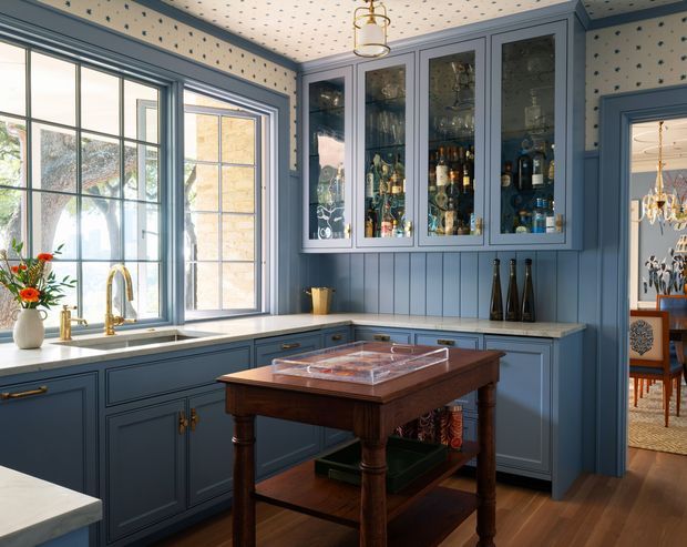 A compact, traditionally styled home bar or entertaining kitchen with deep blue painted cabinetry, a long pale stone benchtop, glass-fronted upper cabinets displaying bottles and glassware, a farmhouse-style sink beneath large divided-pane windows, and a central dark timber island or serving table. The room has blue beadboard wall panelling, patterned wallpaper and a decorative patterned ceiling, with a glimpse of an adjoining dining area through the doorway.